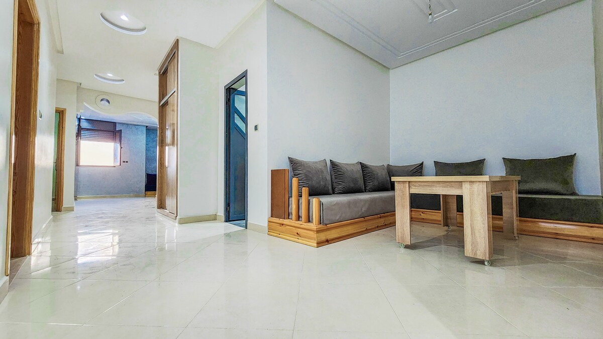 A spacious living area features a wooden coffee table surrounded by a large sectional sofa with plush gray cushions. Natural light enters through a nearby window, illuminating the polished floor tiles, while the entryway is accessed through an open door at the far end.