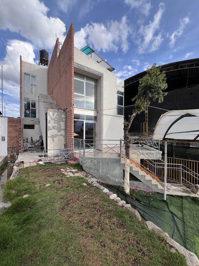 A modern two-story house features large glass windows that provide ample natural light. A sloped pathway leads to the entrance, bordered by lush green grass. The surrounding area includes a large structure, emphasizing the spaciousness of the property.