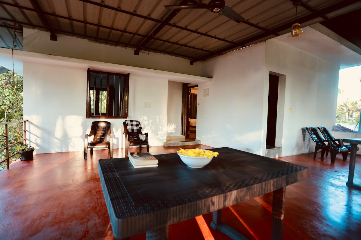 A spacious outdoor area features a dark table with a bowl of yellow fruit at its center. Several black chairs are arranged around the table, and a textured wall serves as a backdrop. Natural light filters in, enhancing the open-air ambiance.
