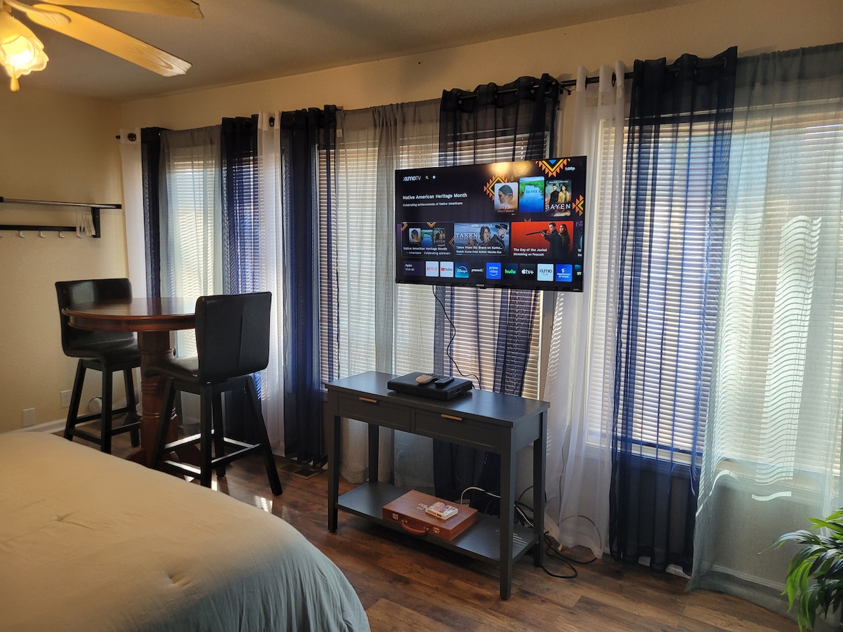 A neutral-toned room features a spacious bed in the foreground. A flat-screen television is mounted on the wall above a low console table, displaying a variety of content. Behind, light-filtering curtains frame the large windows, and a small dining area with two high-backed chairs is visible.