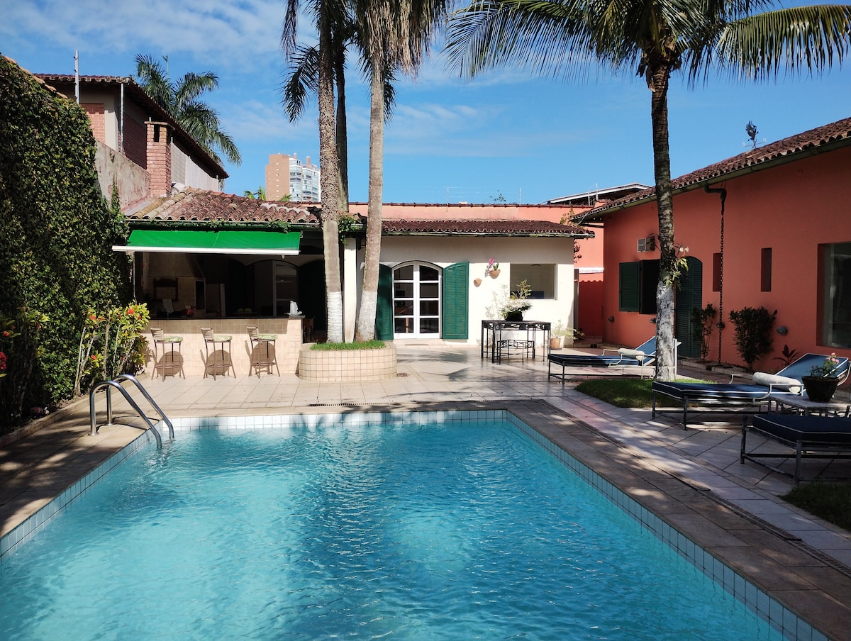 A serene outdoor area features a large swimming pool surrounded by landscaped gardens. Sun loungers line the poolside, and a covered barbecue area with dining tables is visible in the background. The setting is complemented by palm trees and clear blue skies.