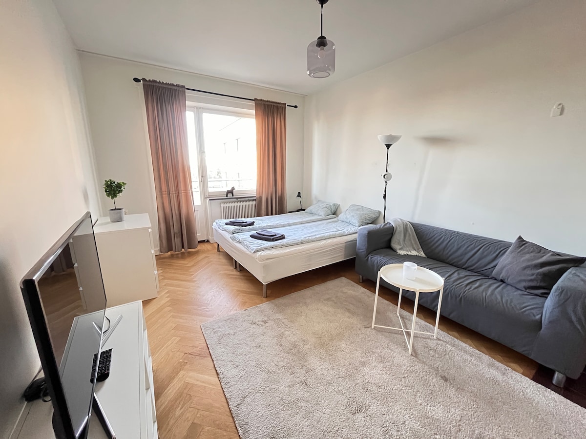 A bright room features a spacious bed with bedding arranged neatly at the foot. A sofa in a soft grey hue provides seating. A small table stands beside the sofa. Large windows are dressed with sheer and heavy curtains, allowing soft natural light to filter in.