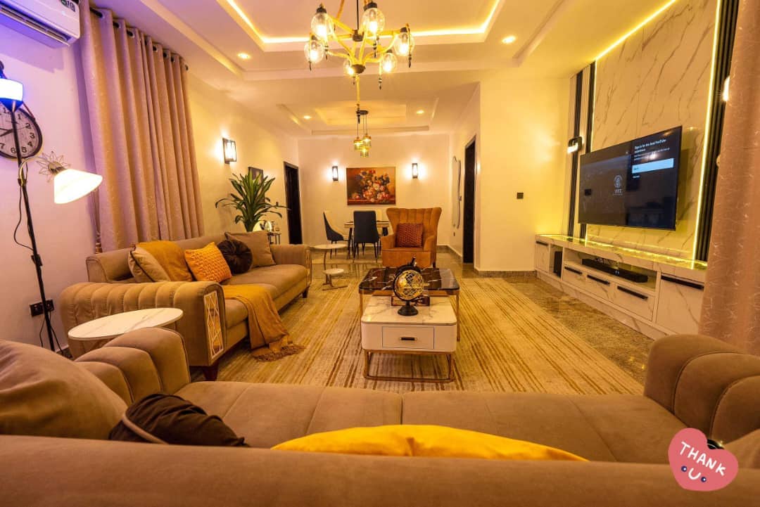 A spacious living area is highlighted by soft lighting and contemporary furnishings. Two comfortable sofas face a sleek media console with a large television. A dining table is positioned in the background, accompanied by stylish chairs. Decorative elements include a plant and tasteful artwork.