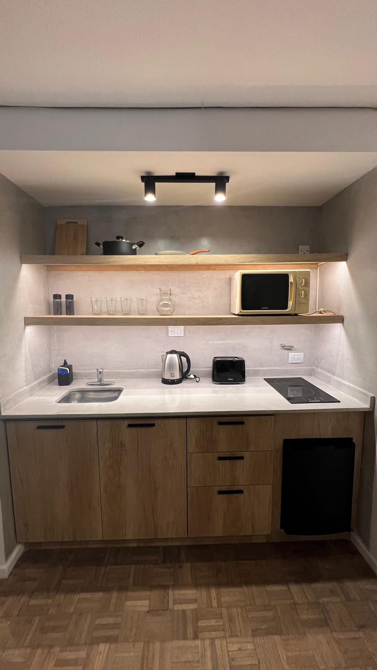 A compact kitchen area features wooden cabinetry and quartz countertops. Small appliances, including a kettle and microwave, are arranged neatly. Above, open shelves hold dishes and a few decorative items, while under-cabinet lighting enhances visibility.