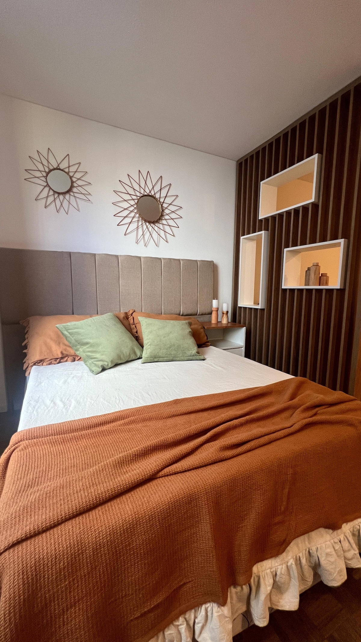 A comfortable sleeping area features a spacious bed with a neutral-coloured blanket draped over it. Two decorative green pillows enhance the bedding, and the walls are adorned with round mirrors. Fitted shelves display a few decorative items, adding a touch of style.