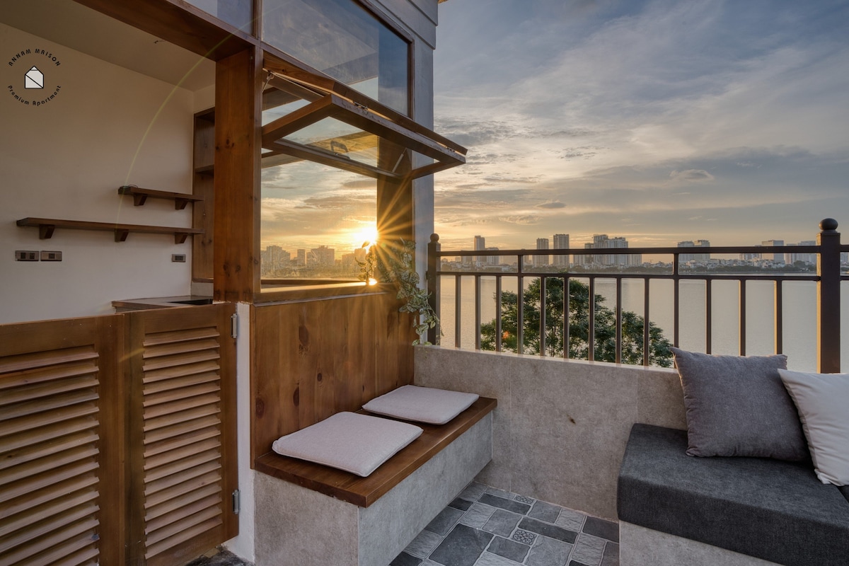 A cozy seating area is positioned by large windows, offering a panoramic view of the city and lake at sunset. Cushions are arranged on a wooden bench, while natural light illuminates the space, enhancing the peaceful ambiance of the setting.