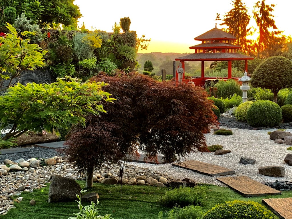 A serene Japanese garden is featured, with lush greenery and carefully arranged stones. A vibrant red pagoda stands prominently at sunset, creating a peaceful scene. Softly textured shrubs and smooth pathways guide the eye through the garden's tranquil layout.