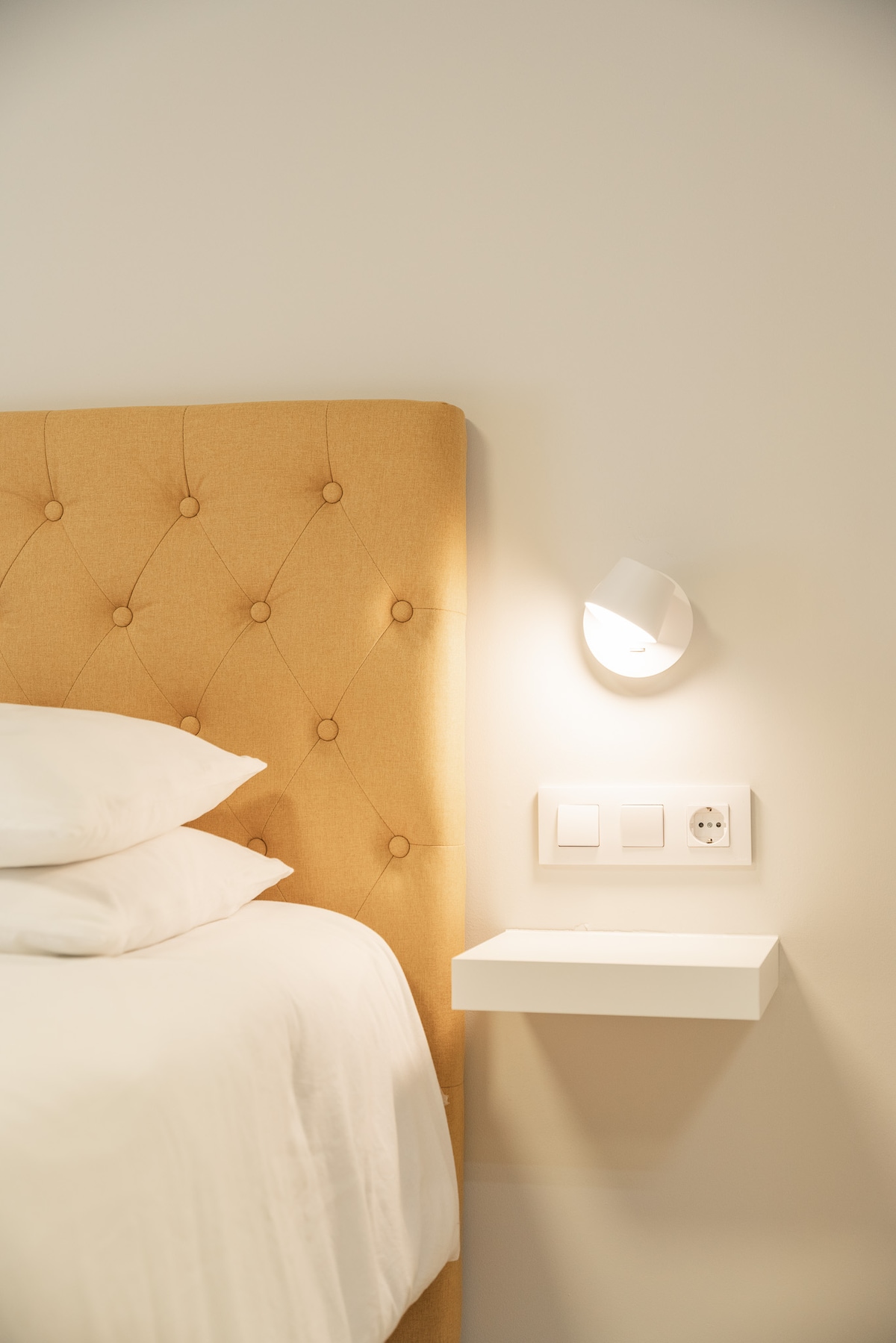 A comfortable sleeping area is highlighted by a soft, tufted headboard in a warm tan shade. A small white bedside table is positioned next to the bed, featuring a wall-mounted lamp and two electrical outlets for convenience.