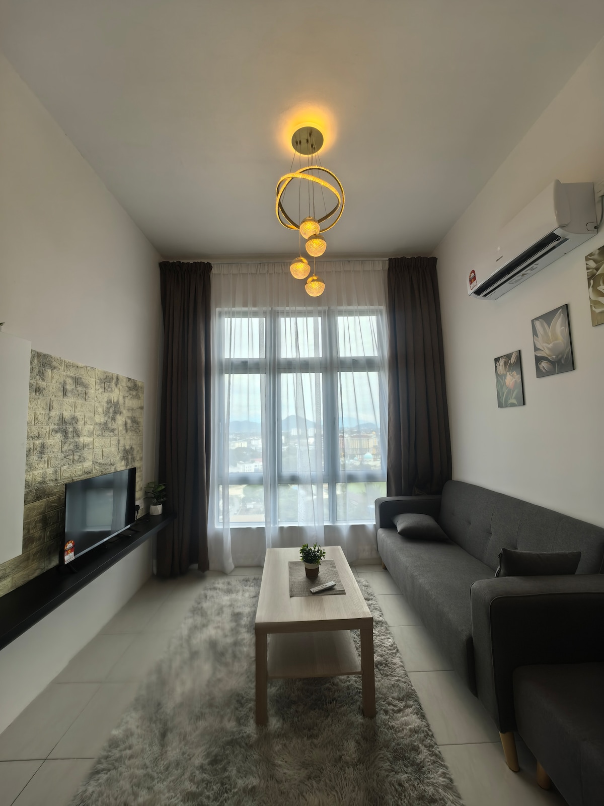 A modern living area is depicted, featuring a comfortable gray sectional sofa and a light wood coffee table placed on a soft area rug. Large windows allow natural light to fill the space, complemented by light curtains. A wall-mounted TV is visible on the opposite wall.