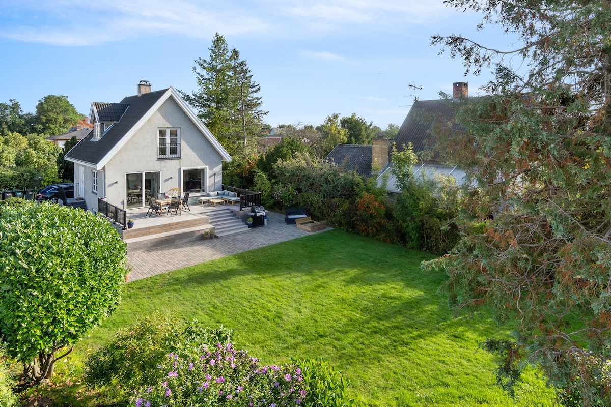 The exterior view showcases a modern house with a spacious terrace and a well-maintained lawn. Seating areas are present on the terrace, surrounded by greenery and flowers, providing a serene outdoor space. The design reflects a harmony with the surrounding natural environment.