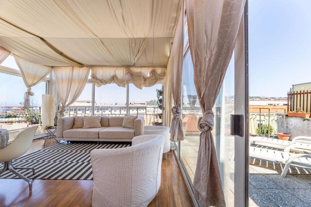 The spacious living area is adorned with soft drapery and large glass windows that provide abundant natural light. A comfortable white sofa faces a contemporary coffee table, while a striped rug enhances the warmth of the wooden flooring. Outside, a balcony can be seen.