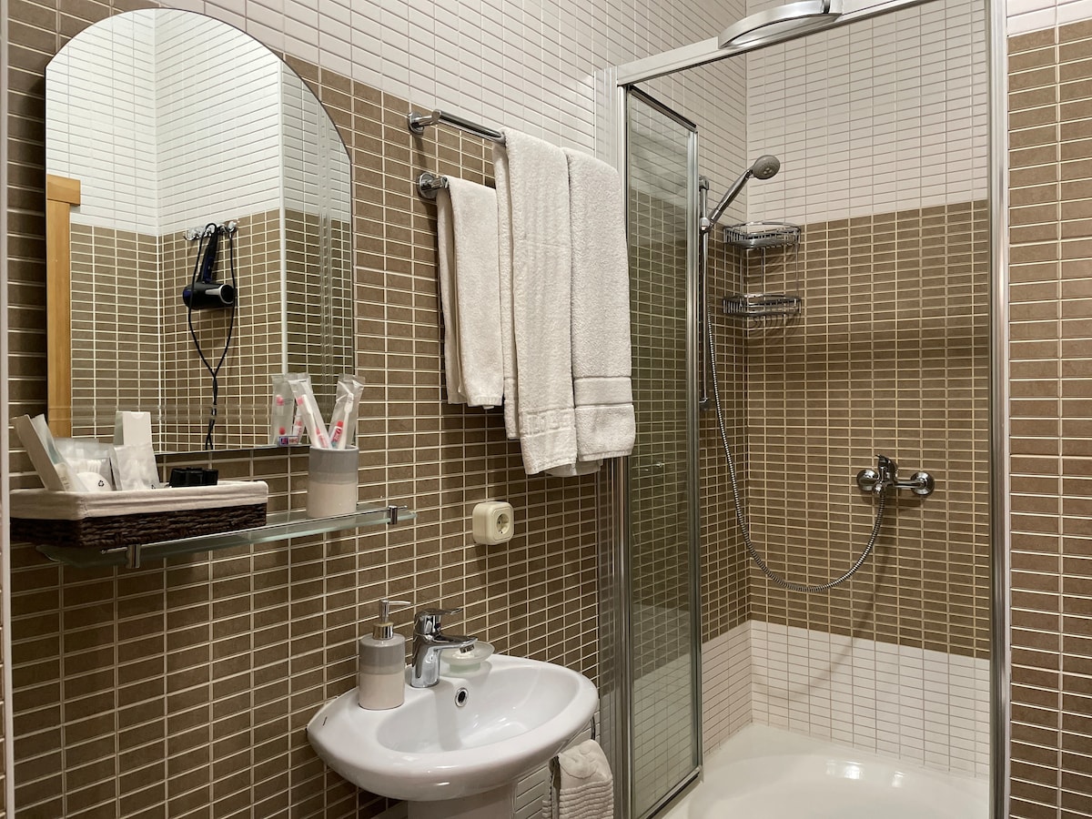 A modern bathroom is shown, featuring a glass shower enclosure and tiled walls in warm tones. Towels are neatly hung on a rack. A round mirror is positioned above a small sink, equipped with toiletries arranged on a shelf.