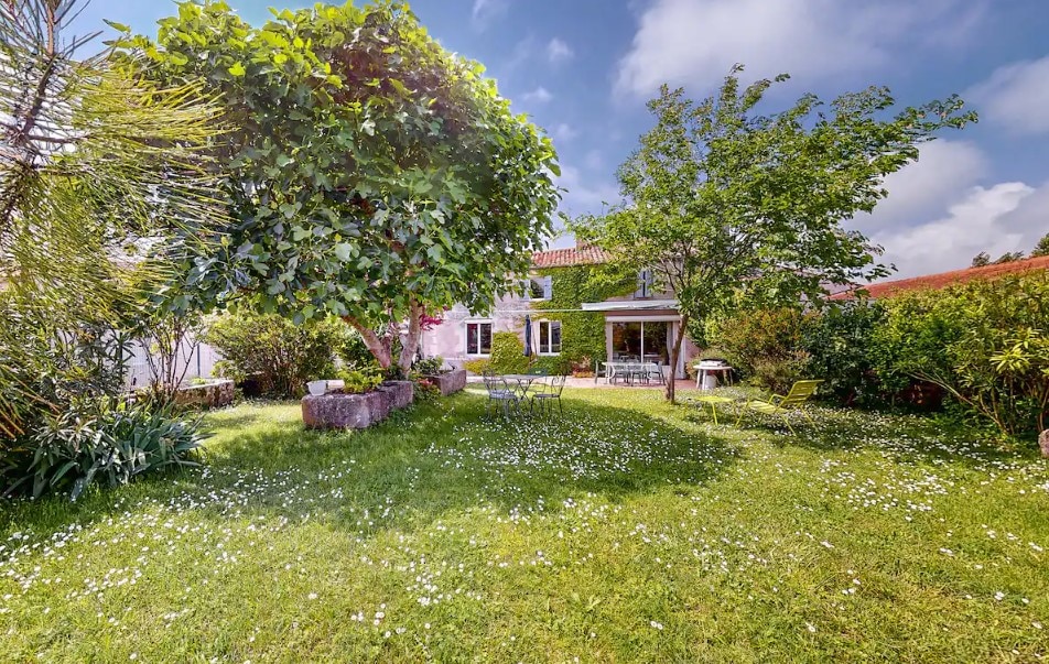 A spacious garden is showcased, featuring lush green grass dotted with white flowers. Two trees provide shade, while garden furniture is arranged for relaxation. In the background, the house is partially obscured by foliage, offering a serene outdoor setting.