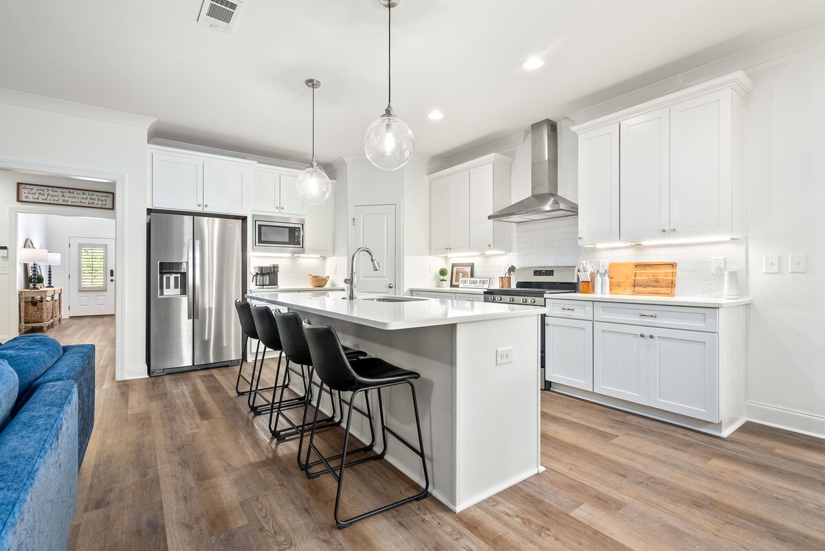 A modern kitchen is showcased with white cabinetry and sleek stainless steel appliances. A large island with seating for four features soft lighting from stylish pendant fixtures. Natural light fills the space from nearby windows, enhancing the open layout.