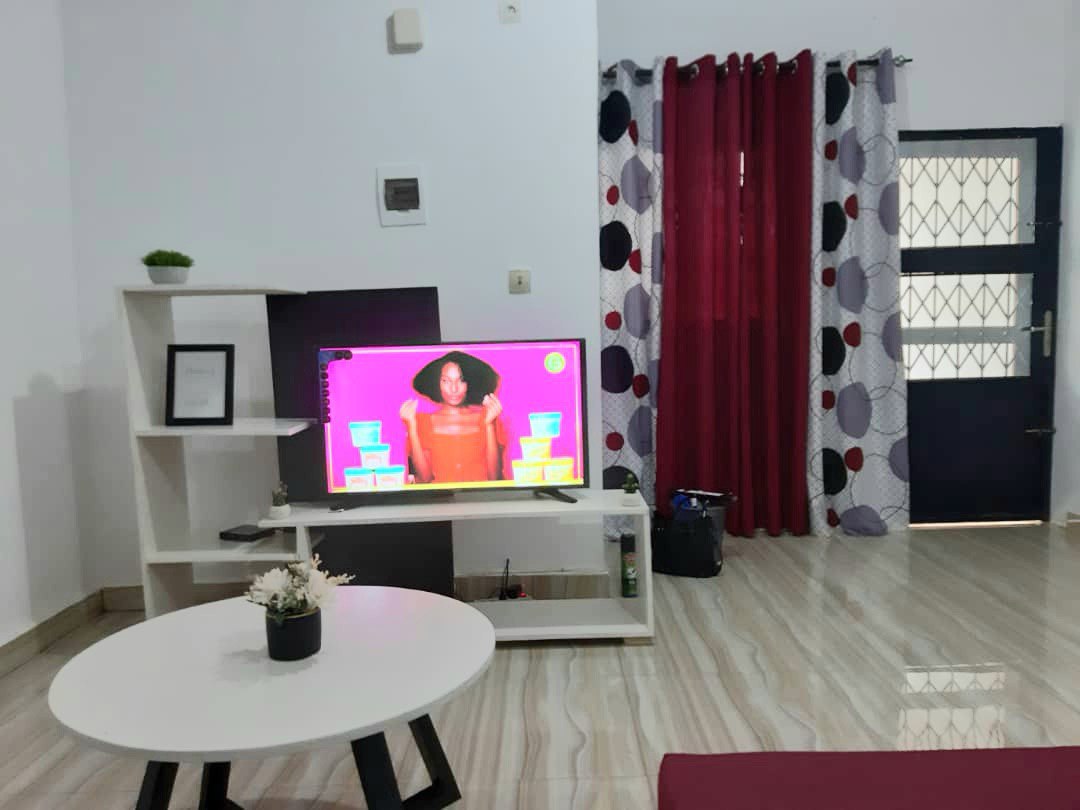 A modern living space is illustrated with a low white coffee table and a sleek entertainment unit featuring a television. Bright red curtains frame the window, and decorative elements are positioned on the shelf and table. The polished flooring adds to the overall clean aesthetic.