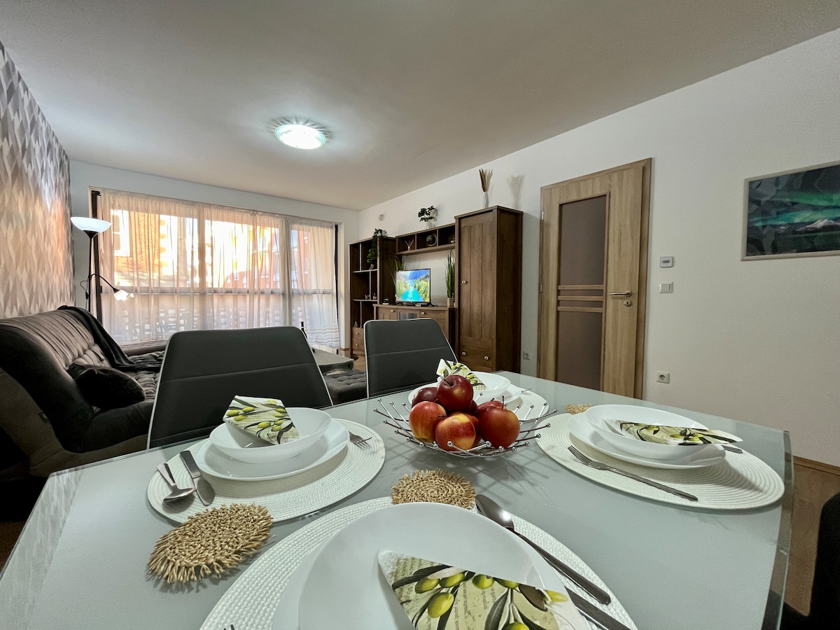 A modern living space is featured, with a dining table set for four, complete with elegant tableware and a bowl of fresh apples. A cozy sofa area is visible in the background, complemented by natural light streaming through large windows.