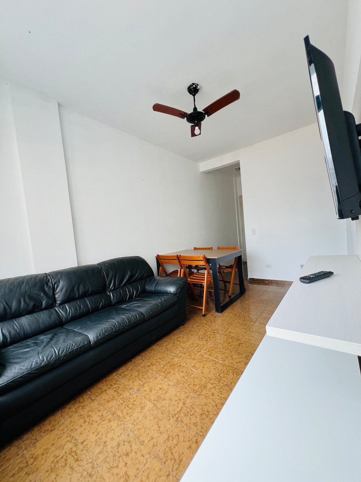 A living area includes a black leather sofa accompanied by a wooden dining table with four folding chairs. A ceiling fan is mounted above, providing air circulation. A wall-mounted television is positioned for easy viewing. Natural light brightens the neutral-toned space.