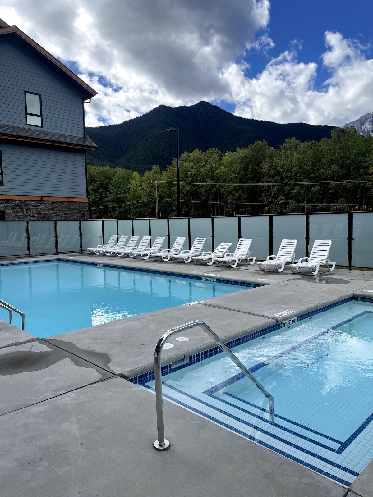 A heated outdoor pool and adjoining hot tub are presented, surrounded by lounge chairs arranged along the deck. The backdrop features rolling mountains under a partly cloudy sky, framed by green trees, creating a relaxing atmosphere.