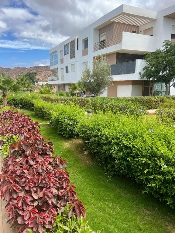 A modern multi-story building is surrounded by lush green landscaping and colorful shrubbery. Palm trees provide a tropical feel, while the sky is partly cloudy, showcasing a mix of blue and white. The soft lawn encourages relaxation in the tranquil outdoor setting.