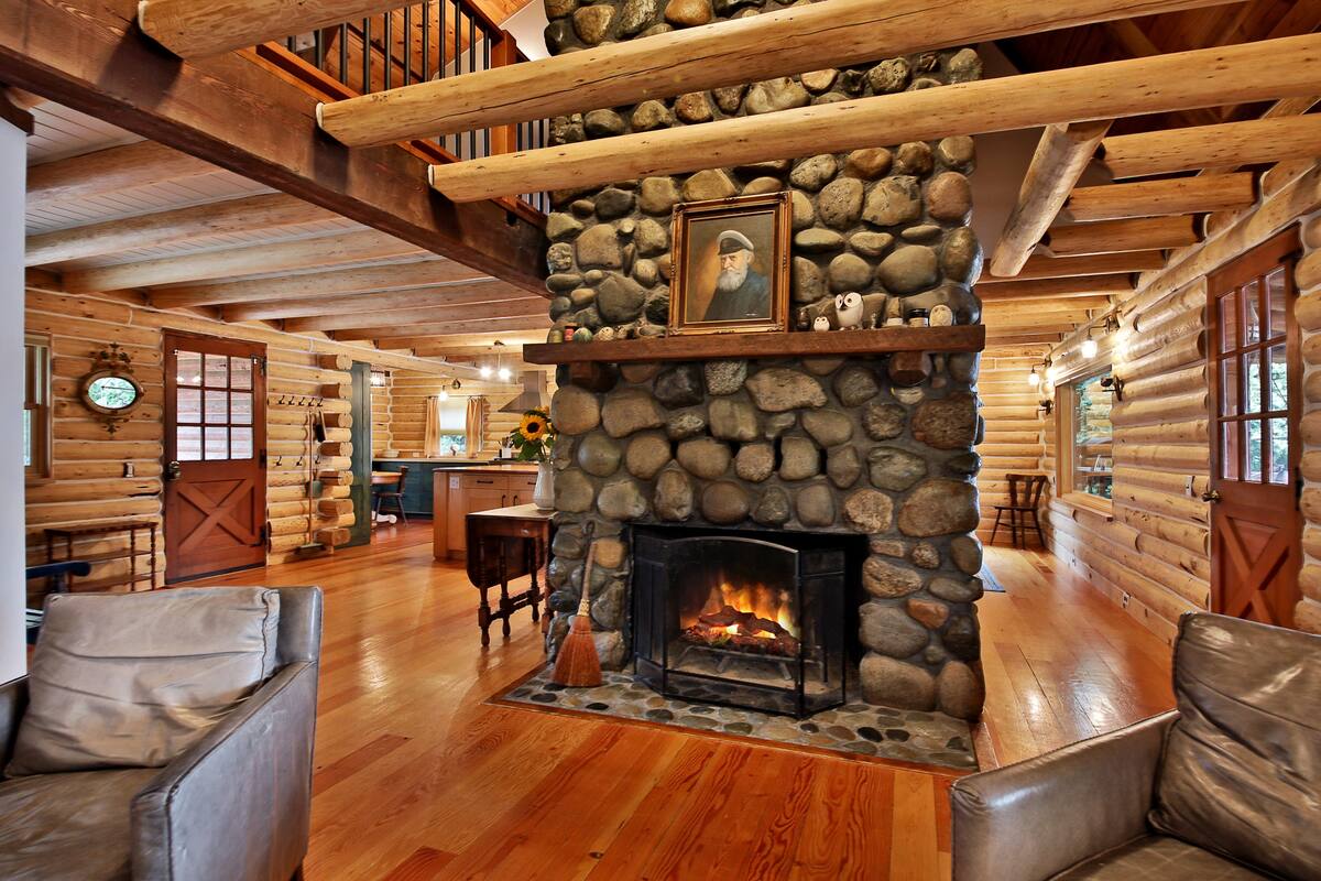 An inviting living area features a large stone fireplace as its focal point, surrounded by warm wooden beams and flooring. Comfortable chairs are positioned nearby, creating a cozy space for relaxation. Natural light fills the room, highlighting the rustic log walls and charming decor.