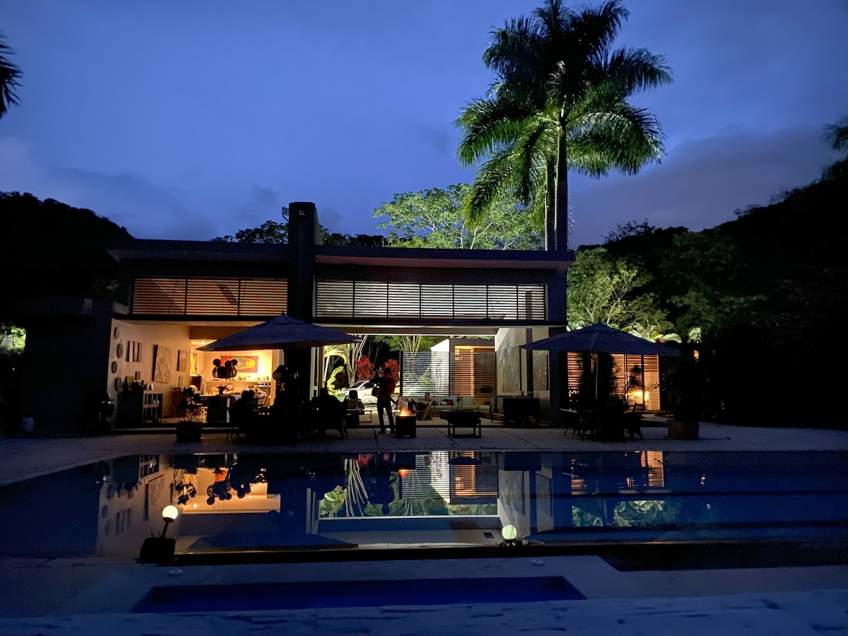 The modern structure features large windows, revealing an inviting interior at dusk. Outdoor seating areas are set under umbrellas, and the tranquil pool reflects the surrounding greenery and illuminated building, creating a serene ambiance.