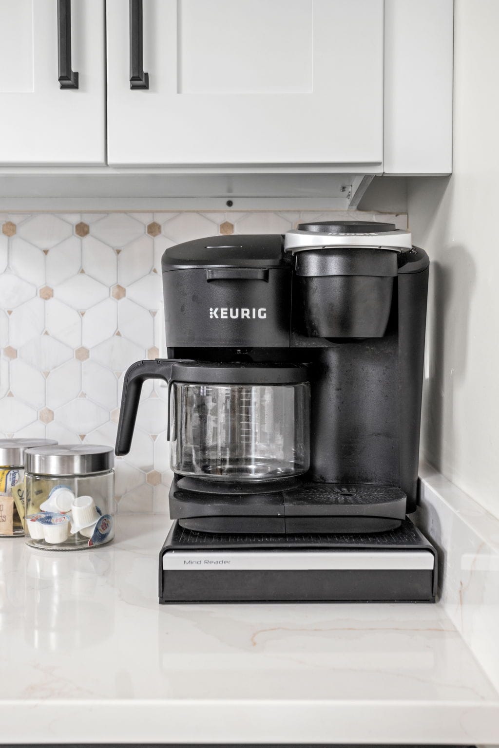 A modern coffee maker sits prominently on a sleek countertop. The appliance features a glass carafe alongside a single-serve brewing option. Behind, neatly arranged coffee pods and storage canisters are visible, enhancing the kitchen's functionality and organization.