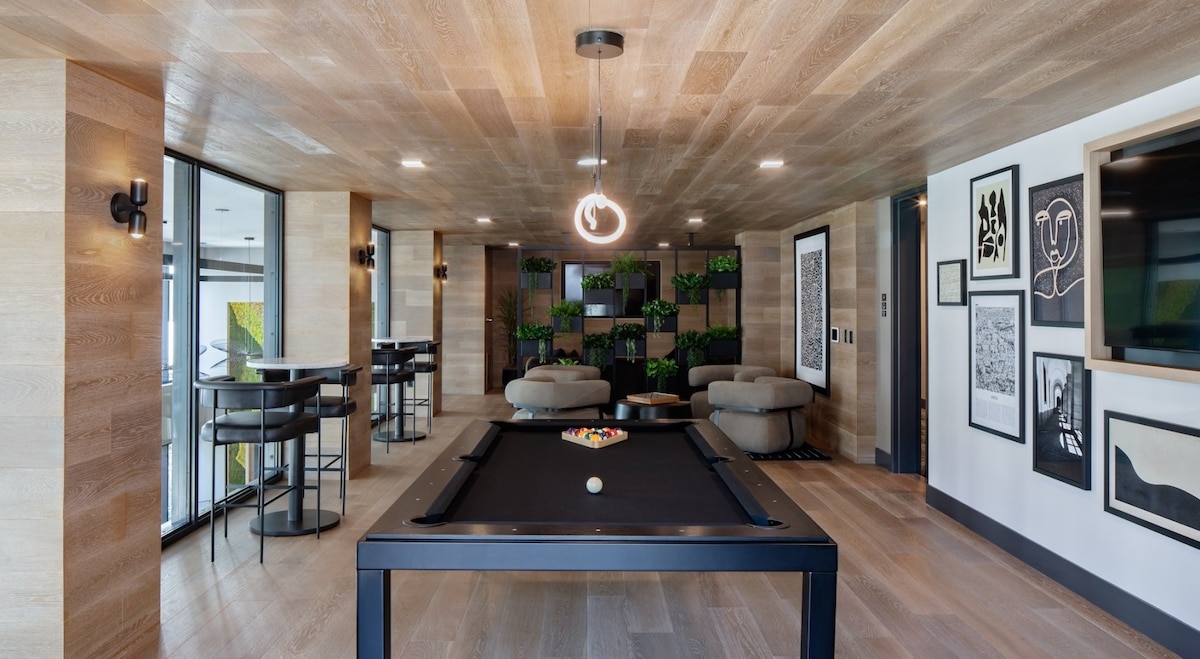 A modern game room features a sleek black pool table centered under a unique light fixture. Comfortable seating is arranged around the room, and large windows offer natural light. Walls are decorated with framed artwork and greenery, creating an inviting social space.