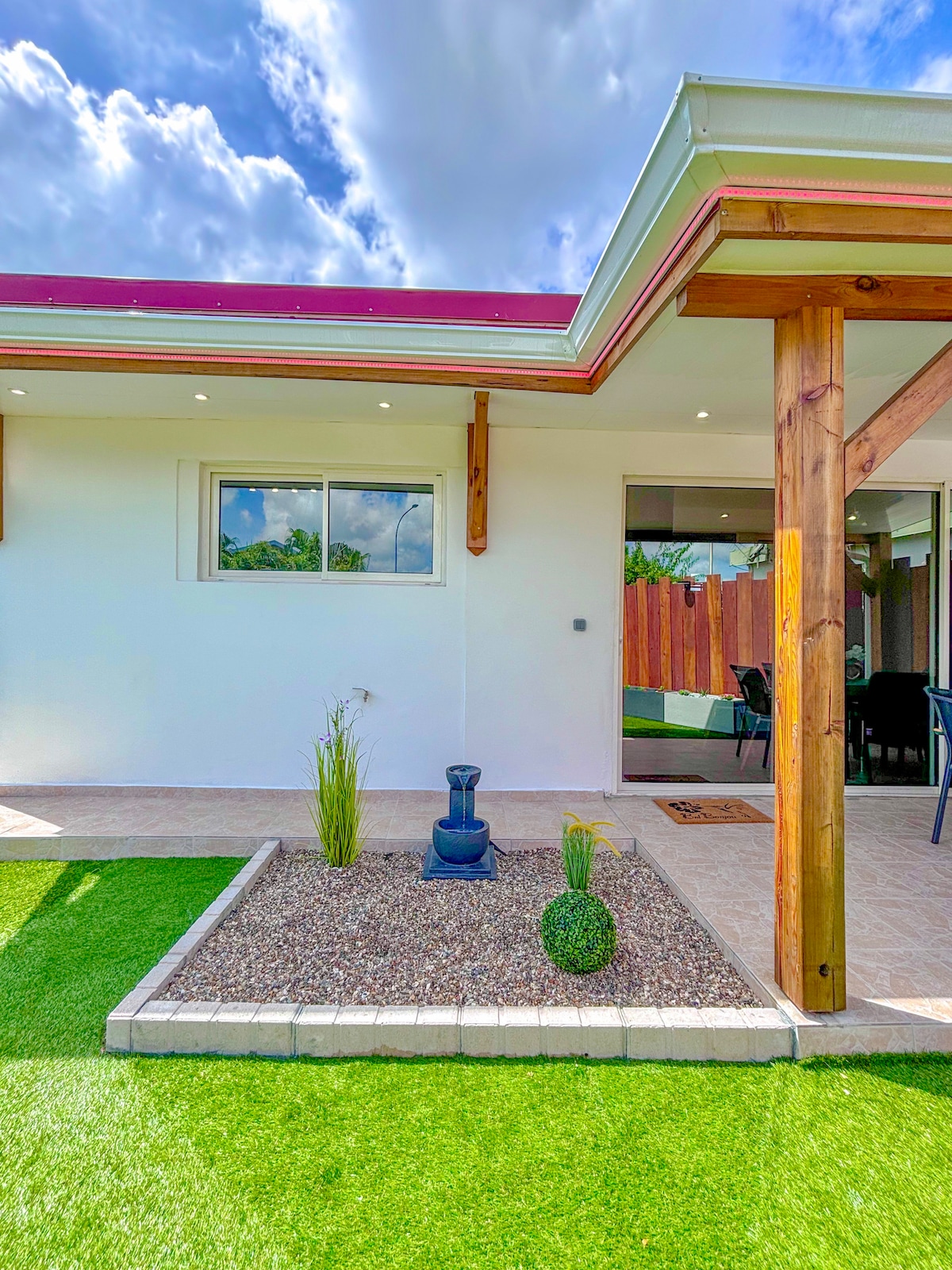 A well-maintained outdoor area features lush green artificial grass and a decorative garden bed containing small plants and stones. A simple water feature is positioned at the center, and a shaded entryway with wooden beams can be seen leading into the studio.