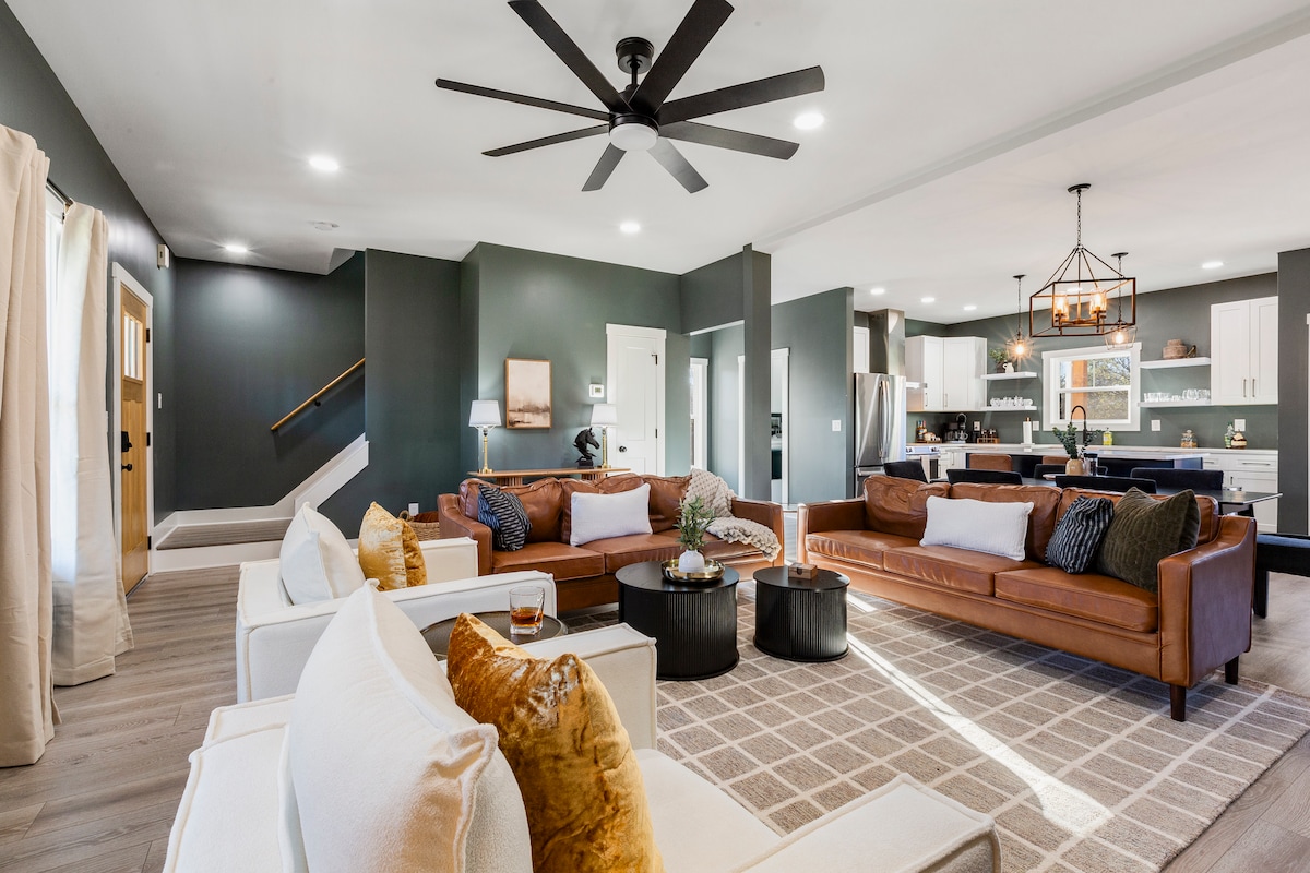 A spacious living room is showcased with two leather sofas and white armchairs arranged around a circular coffee table. A modern ceiling fan hangs above, while a large kitchen is visible in the background, featuring white cabinetry and dark accents. Large windows allow natural light to fill the room.