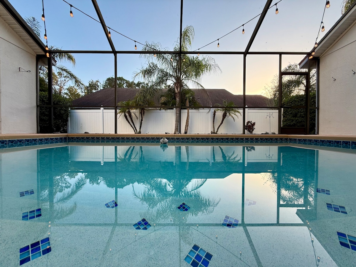 A private pool is situated within a screened enclosure, reflecting the clear blue sky and surrounding greenery. Strings of lights hang above, enhancing the tranquil setting. The water is calm, showcasing tiles at the bottom and providing a serene backdrop for relaxation.
