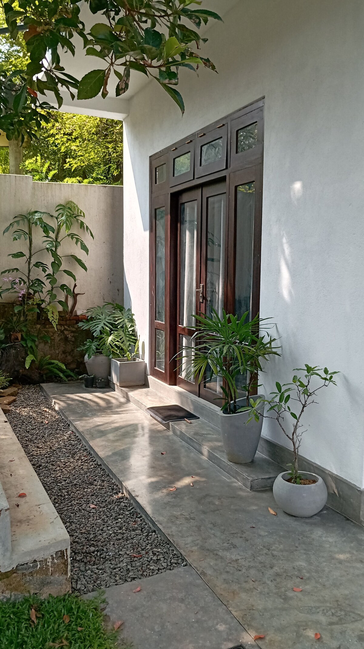 The entrance is framed by two large, wooden doors, flanked by potted plants. A smooth stone path leads to the doorway, with a mix of gravel and greenery creating a natural ambiance. Sunlight gently illuminates the area, enhancing the tranquil atmosphere.