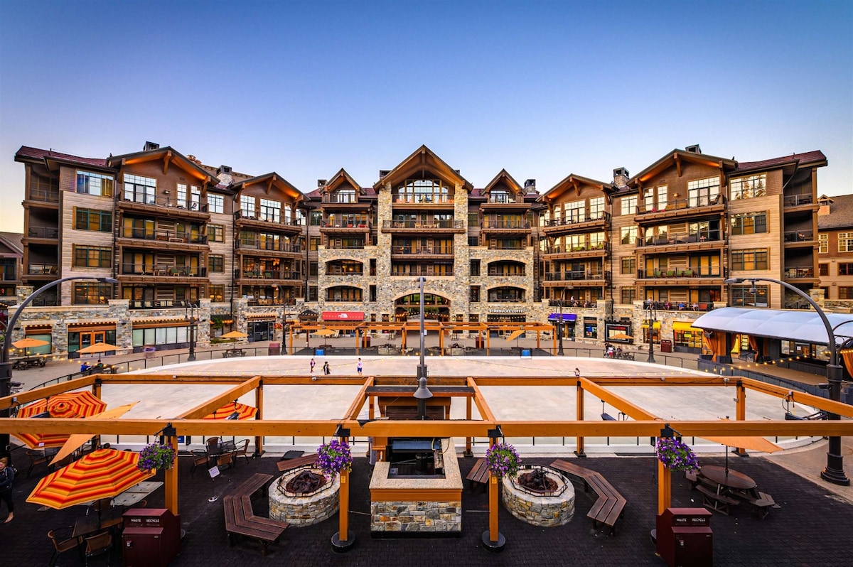 A lively village square is presented with a skating rink at its center, surrounded by charming storefronts featuring wooden and stone facades. Outdoor seating areas are arranged around the rink, with orange umbrellas providing shade. Mountain views serve as a backdrop to this vibrant destination.