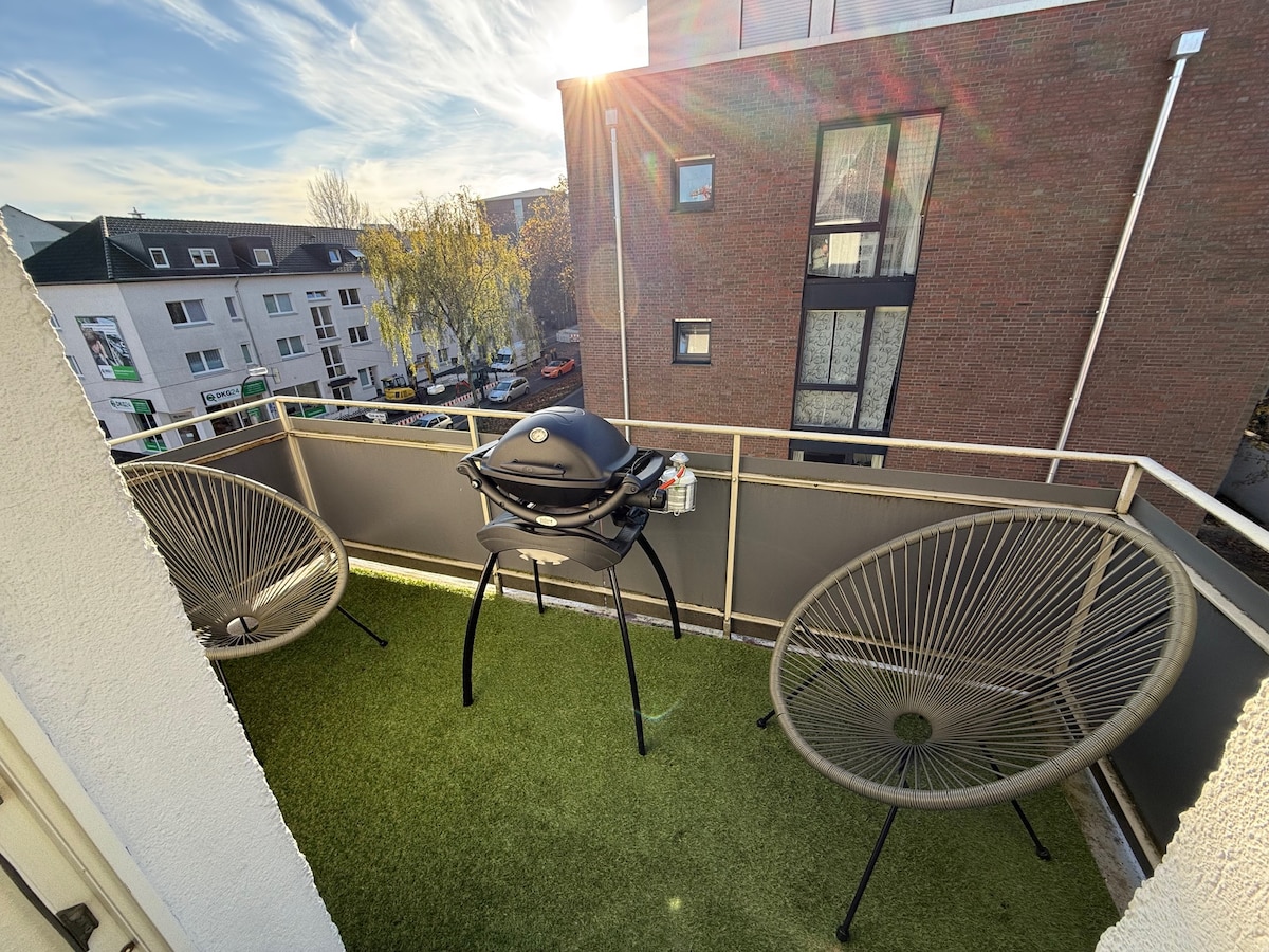 A private balcony is shown, featuring two stylish chairs and a Weber gas grill positioned on green synthetic grass. Sunlight bathes the area, highlighting the surrounding buildings and distant street view. The balcony offers a comfortable outdoor space for relaxation.
