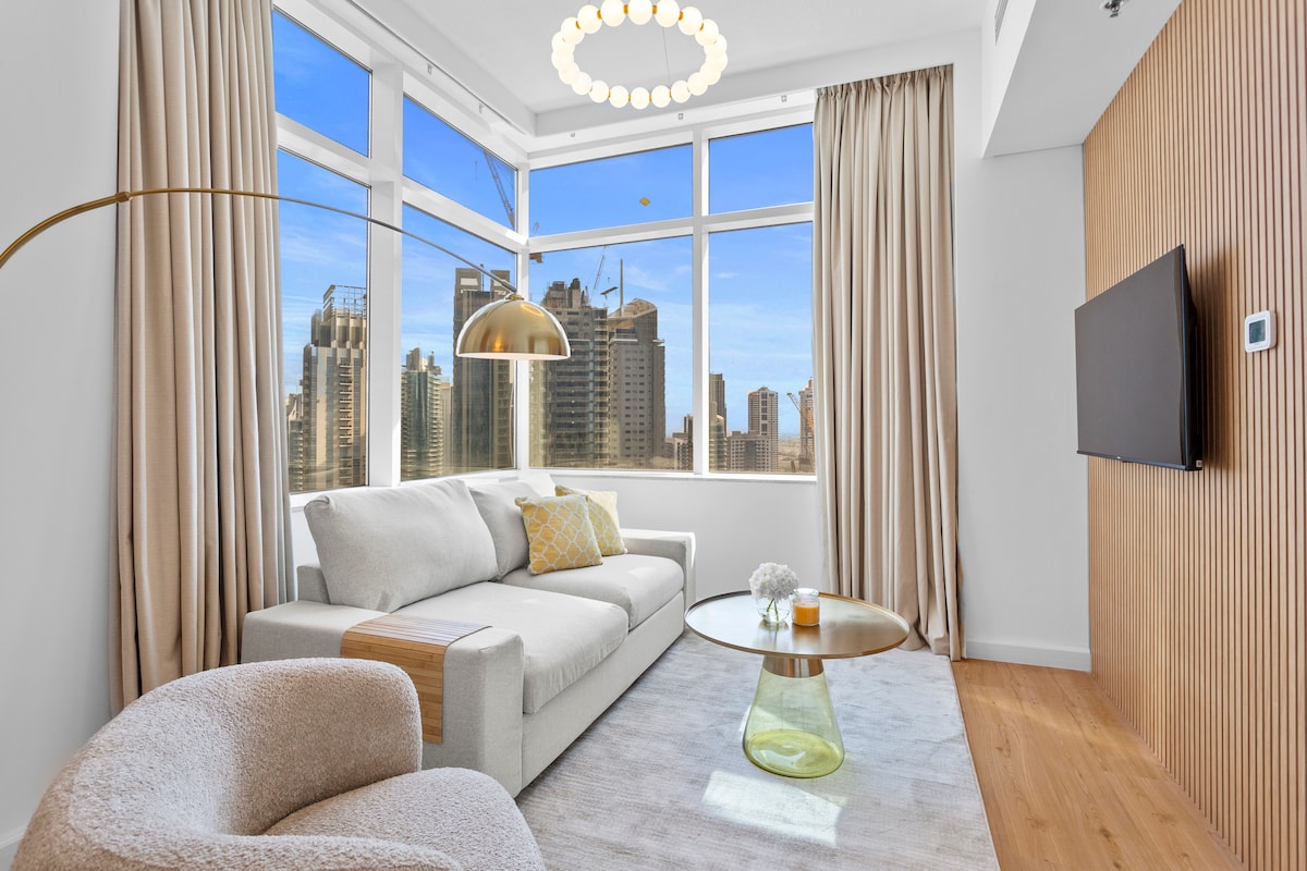 A bright living area features large windows offering city views. A light-colored sofa is complemented by a round glass coffee table, while a plush accent chair provides additional seating. Soft curtains frame the windows, and a modern light fixture hangs overhead, enhancing the contemporary design.