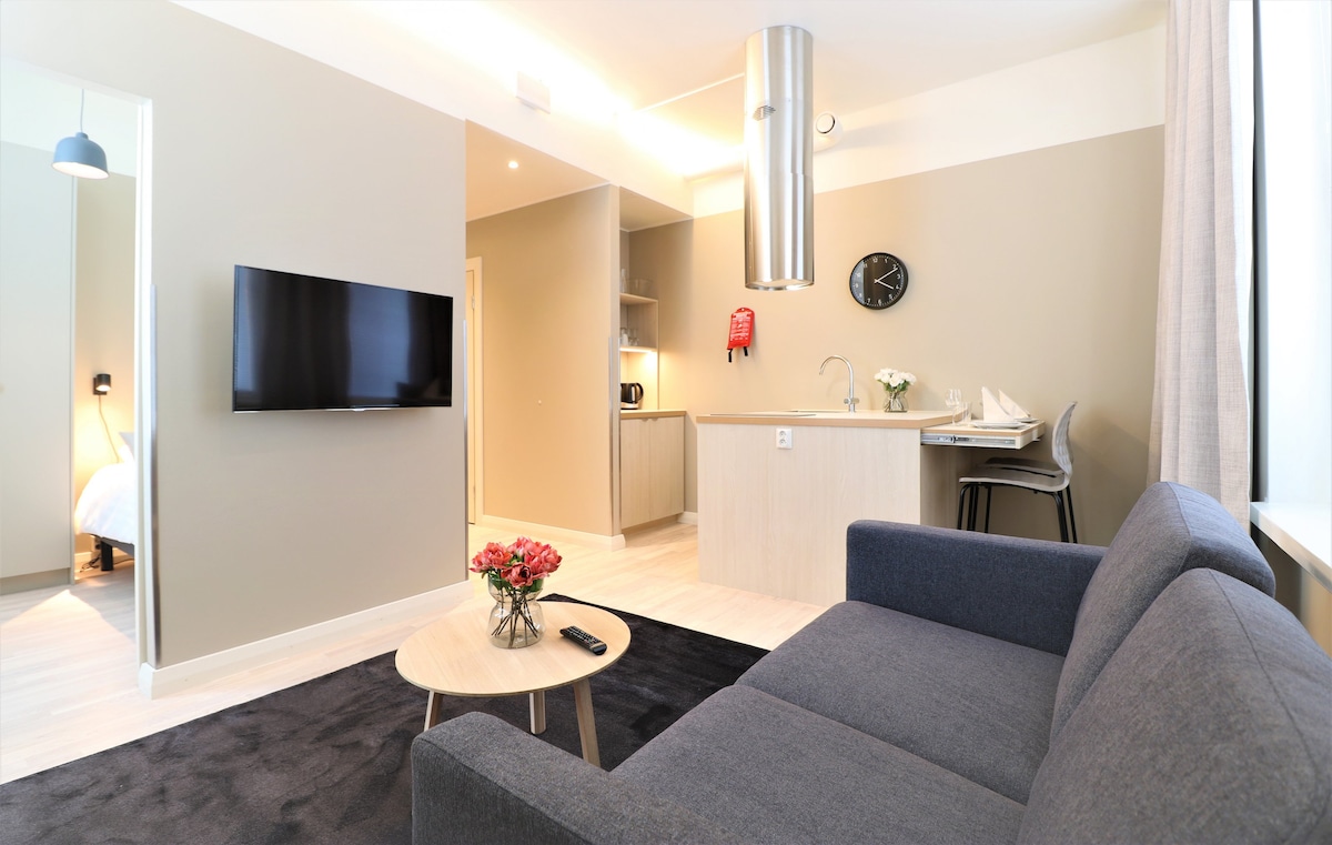 A modern living area features a comfortable gray sofa facing a wall-mounted television. A round coffee table sits in front, and a kitchenette with bar stools is visible in the background. Soft light enters through a nearby window, illuminating the neutral-toned walls.