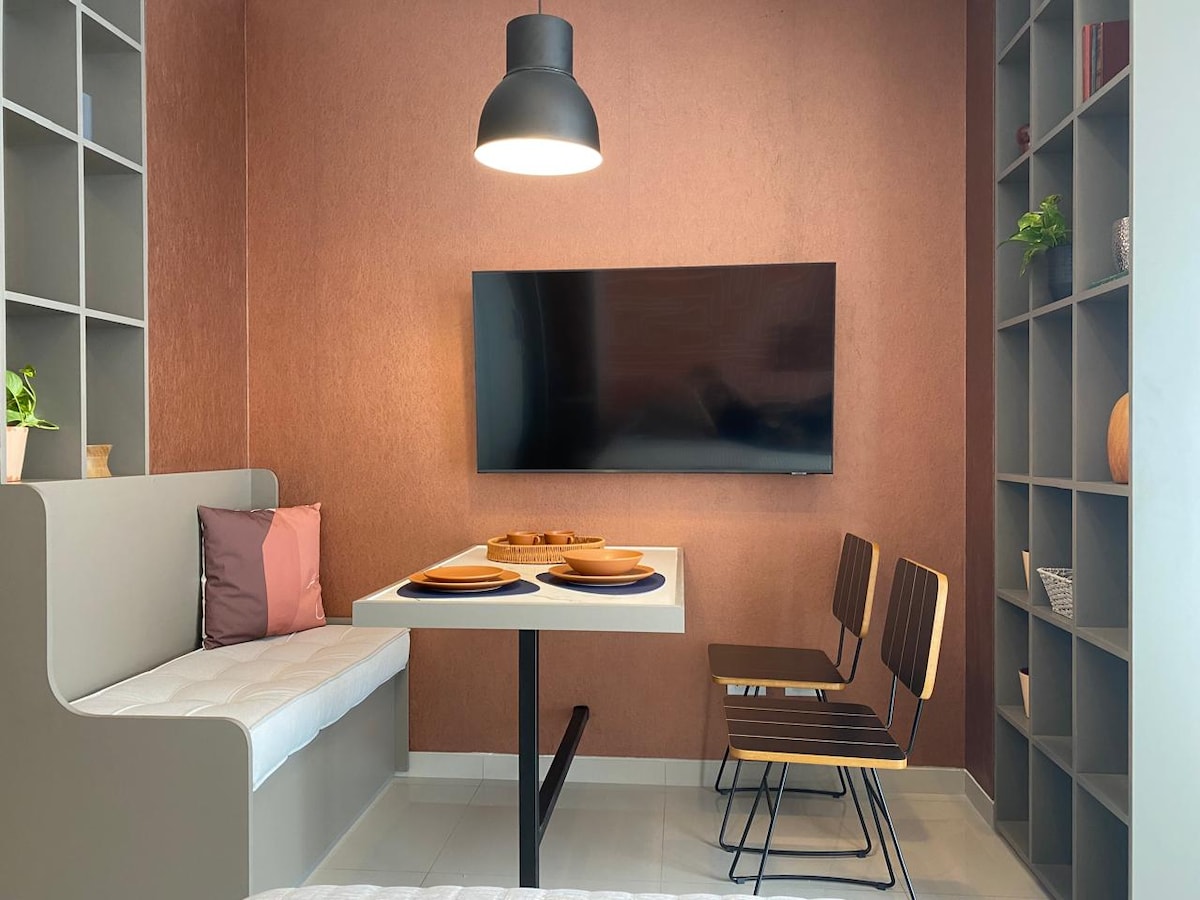 A modern dining area features a sleek white table accompanied by two wooden chairs. A cozy bench with soft cushions is positioned against the wall. A large television is mounted nearby, with warm lighting provided by a hanging pendant lamp, creating an inviting space.