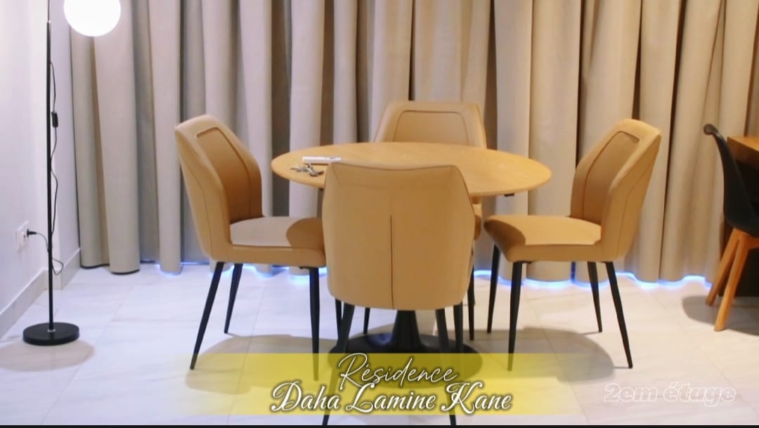 An inviting dining area features a round table with four beige chairs, designed for comfort. Soft curtains frame the space, while ambient lighting creates a serene atmosphere. A floor lamp stands to one side, contributing to the overall warmth of the room.