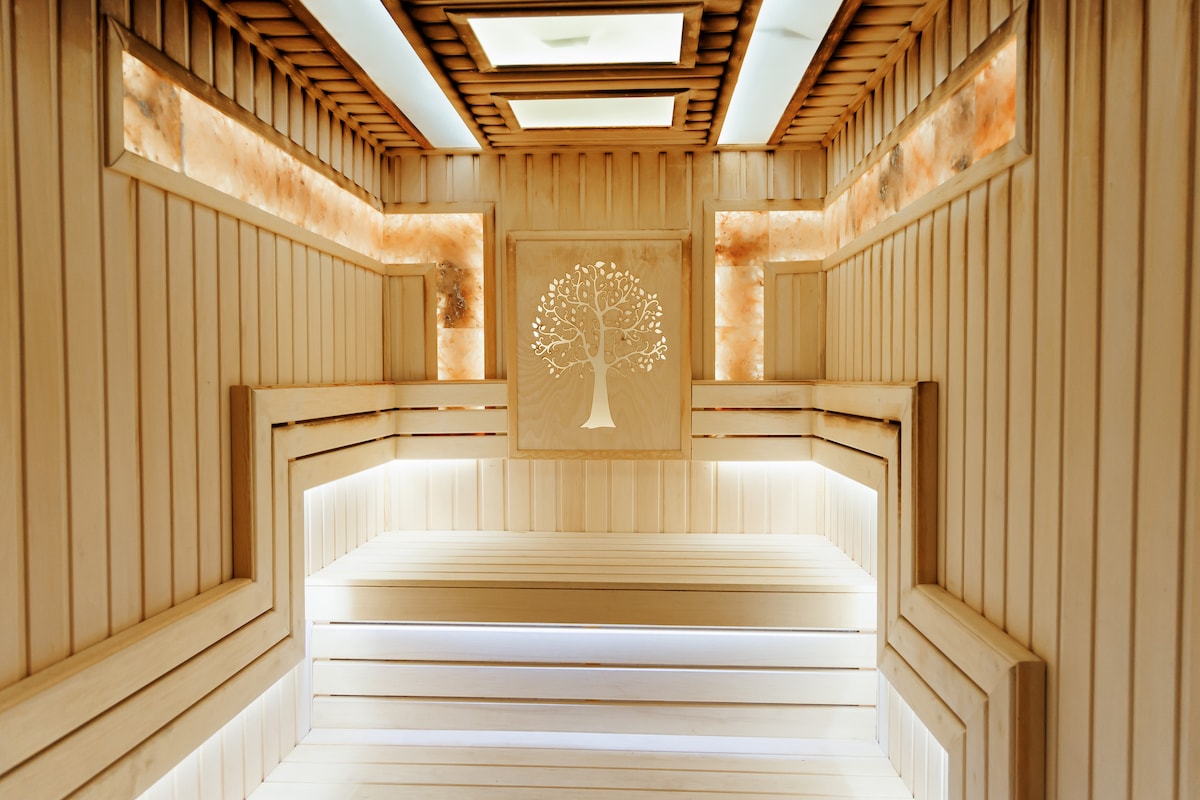 A serene sauna space is presented with smooth wooden benches and paneling. Soft lighting highlights the walls, featuring decorative elements of Himalayan salt and an artistic tree design, creating a calming environment for relaxation.
