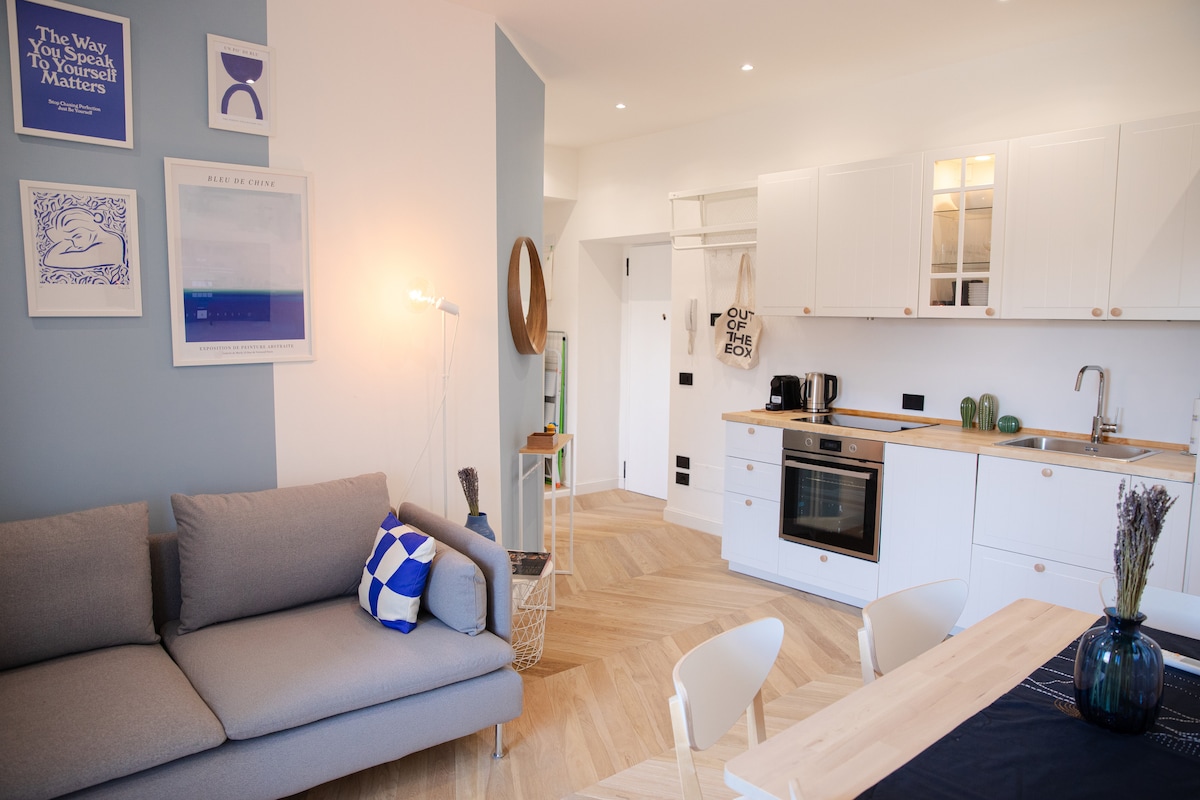 An open-concept living area features modern furnishings, including a light gray sofa adorned with a blue and white cushion. A dining table is visible, surrounded by white chairs. The kitchen is equipped with sleek cabinetry and appliances, complemented by natural wood accents. Natural light brightens the space through the expansive window.