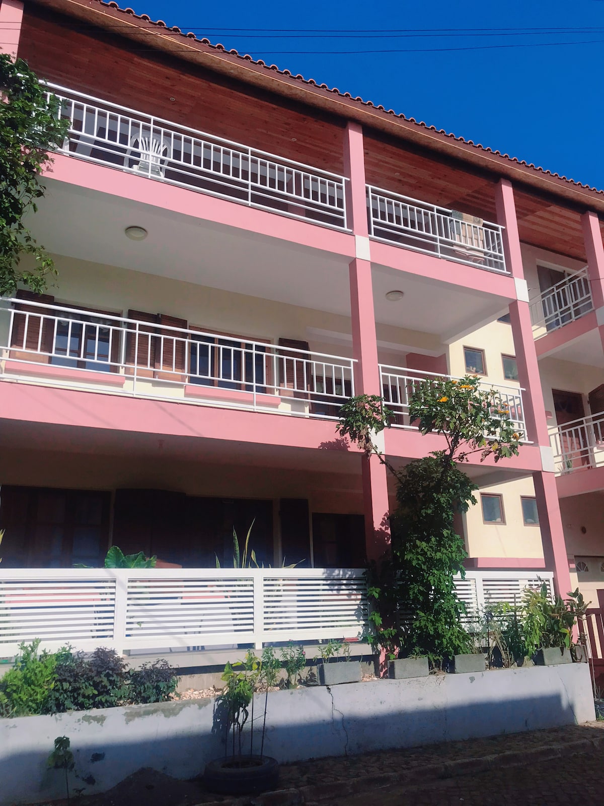 A three-story building is displayed with a light pink exterior and white railings. Lush greenery and plants are visible in the foreground, contributing to the building's inviting appearance. Expansive balconies are seen on each floor, allowing for outdoor relaxation.