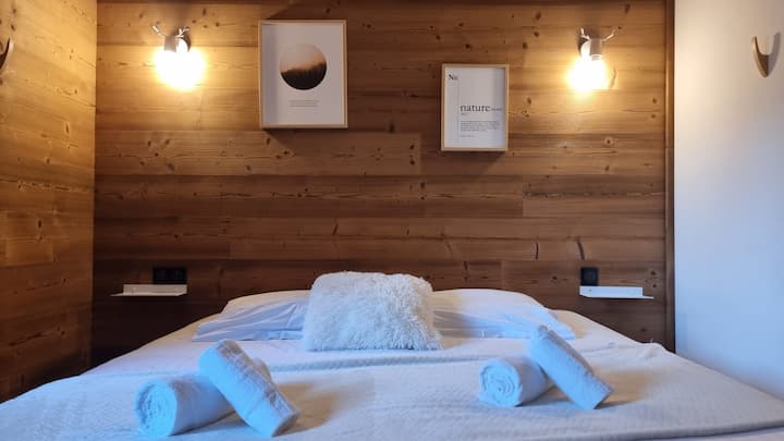 غرفة نوم مريحة في الشاليه الخشبي، مع ملاءات بيضاء نقية وإضاءة ناعمة لمصباحين جداريين، مما يخلق جوًا مريحًا ودافئًا لليالي المريحة.