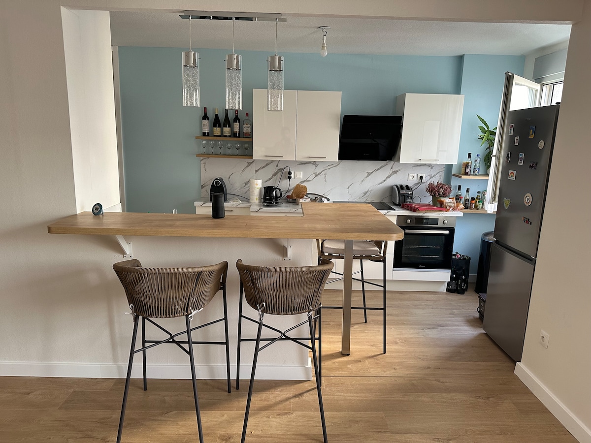 A modern kitchen area features light wood countertops and stylish bar stools. Hanging glass light fixtures provide illumination above the counter. The kitchen is equipped with sleek appliances, including an oven and a refrigerator, with various bottles and kitchenware neatly organized on adjacent shelves.