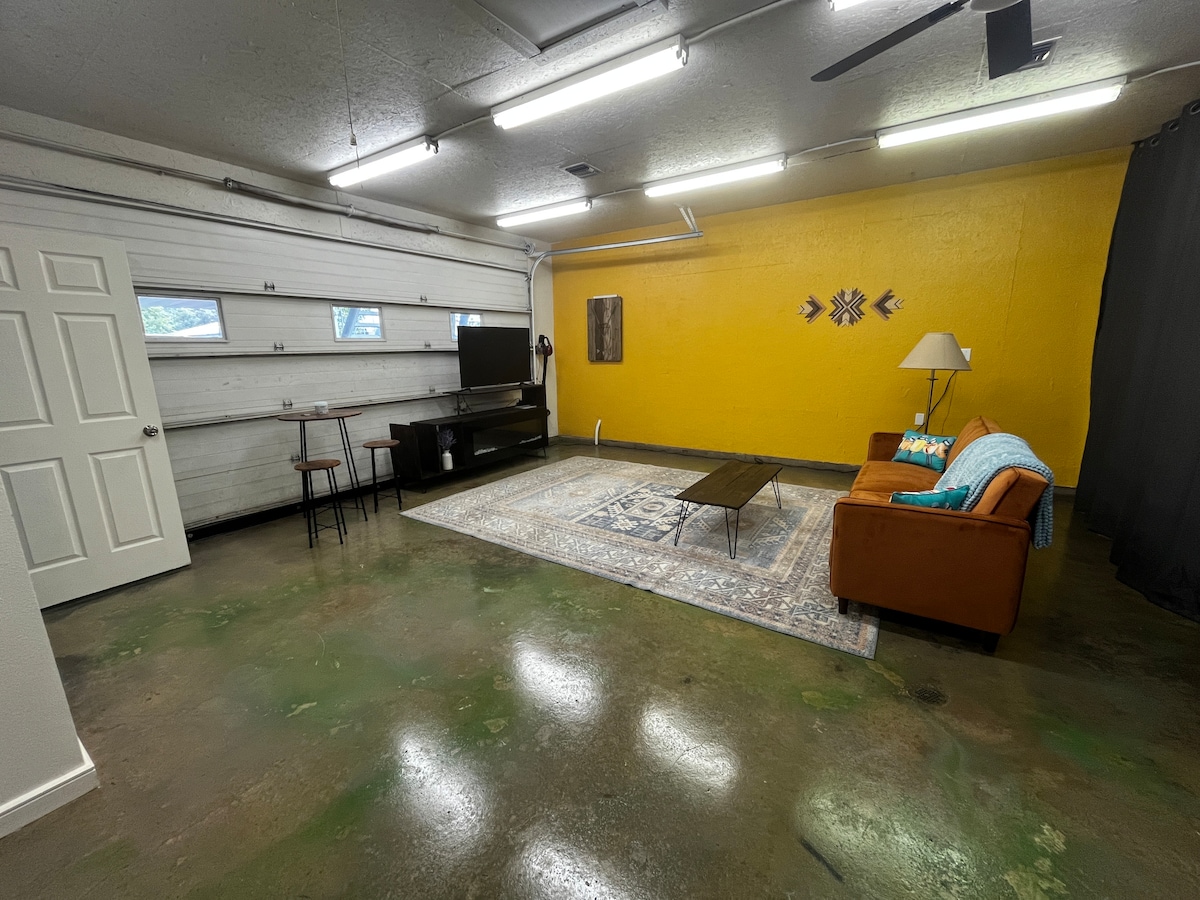 A spacious studio area features a bright yellow wall, providing a vibrant atmosphere. A comfortable brown couch is paired with a side table and lamp. A TV is positioned on a low stand next to a wooden dining table with two chairs. Large garage-style doors allow for ample lighting.