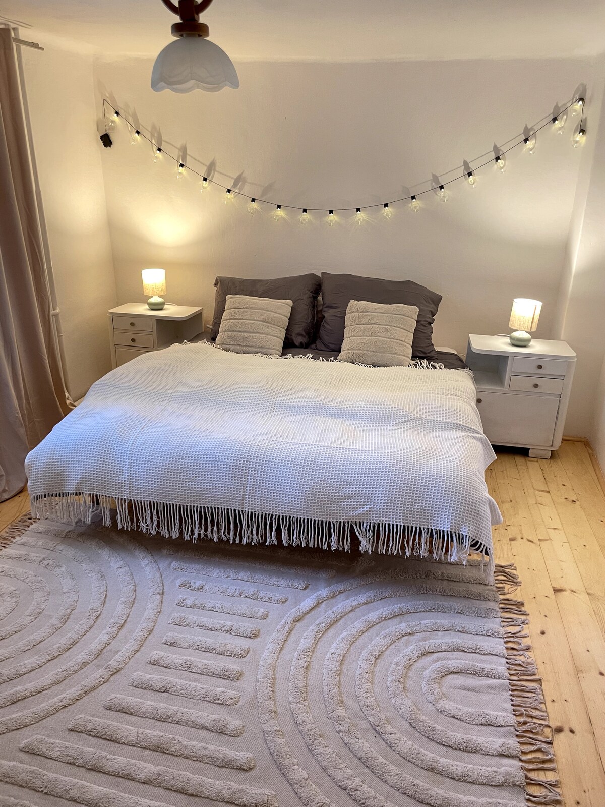 A cozy sleeping area is presented with a spacious bed adorned with a soft white blanket. Two nightstands with lamps provide gentle lighting, while string lights are hung above the bed, adding a touch of warmth. A textured area rug lies underneath, complementing the wooden floor.