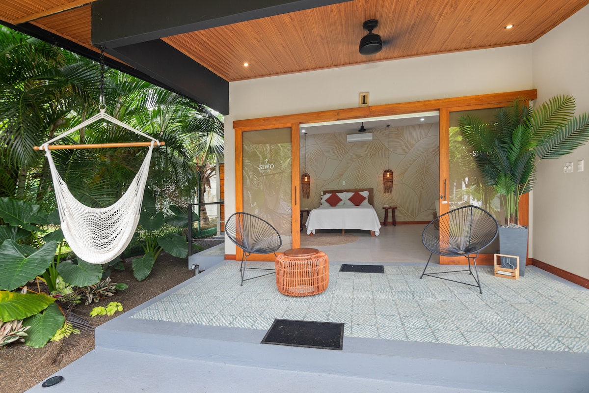 A private terrace is furnished with two dark chairs and a round wicker table, surrounded by lush tropical plants. A hammock is suspended for relaxation. The entrance leads to a well-lit suite with soft tones, enhancing the connection with nature.