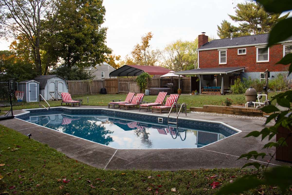 A backyard with a serene pool is surrounded by a grassy lawn and several trees. Lounge chairs with red and white stripes are positioned around the pool. In the background, a covered patio and a utility shed are visible, contributing to a relaxing outdoor space.