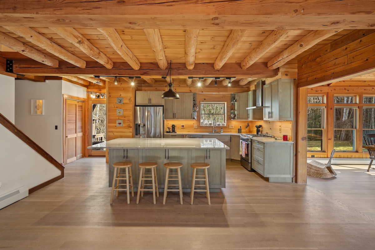 A spacious kitchen features an island with four stools, complemented by wooden beams overhead. The modern design includes stainless steel appliances and ample counter space. Large windows provide natural light, showcasing views of the surrounding woods.