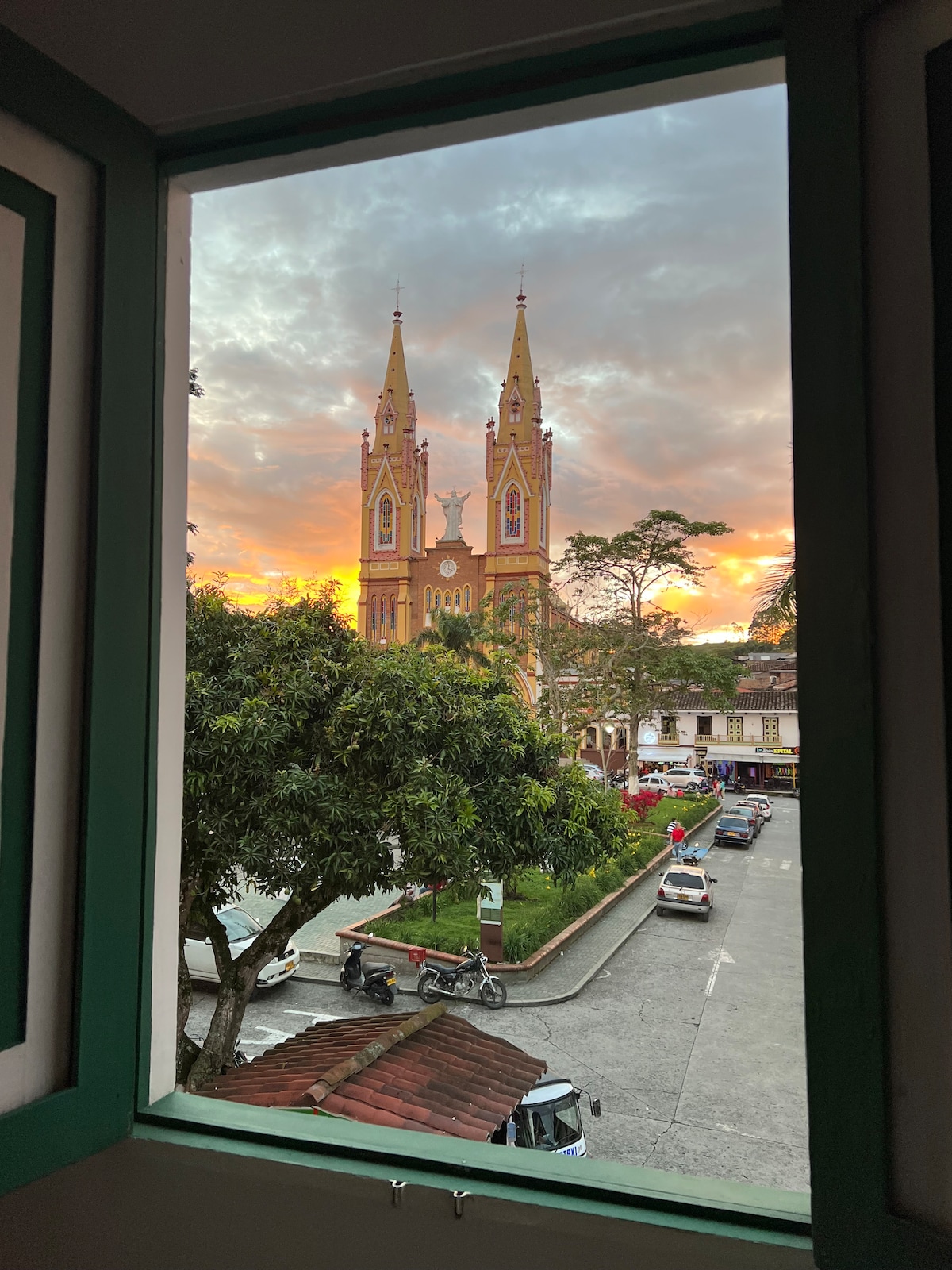 A view is captured through a green-framed window, showcasing the vibrant church with twin spires against a colorful sunset. Trees and buildings are visible in the foreground, with a quiet street lined by parked vehicles.