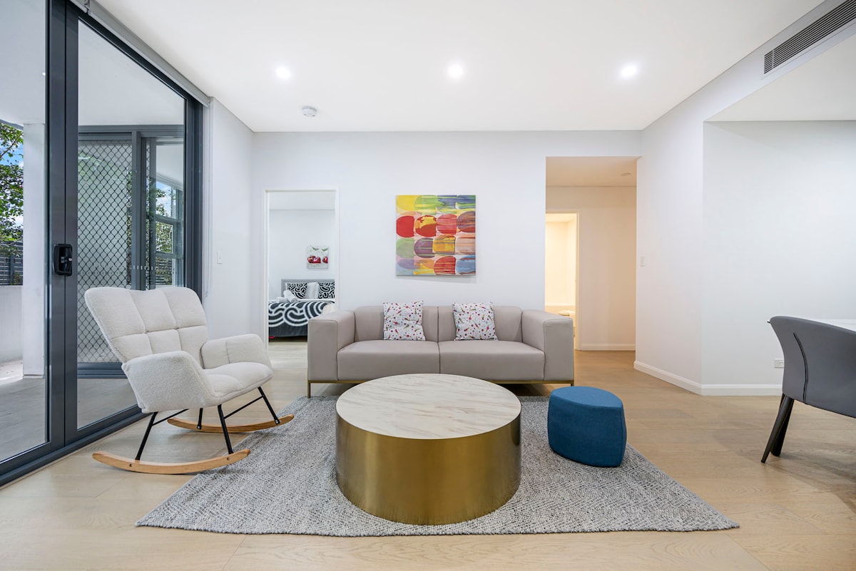 A modern living area features a comfortable sofa positioned across from a round coffee table. Natural light fills the space through large glass doors, which lead to an outdoor area. A colorful abstract painting decorates the wall, complemented by a cozy armchair and ottoman.