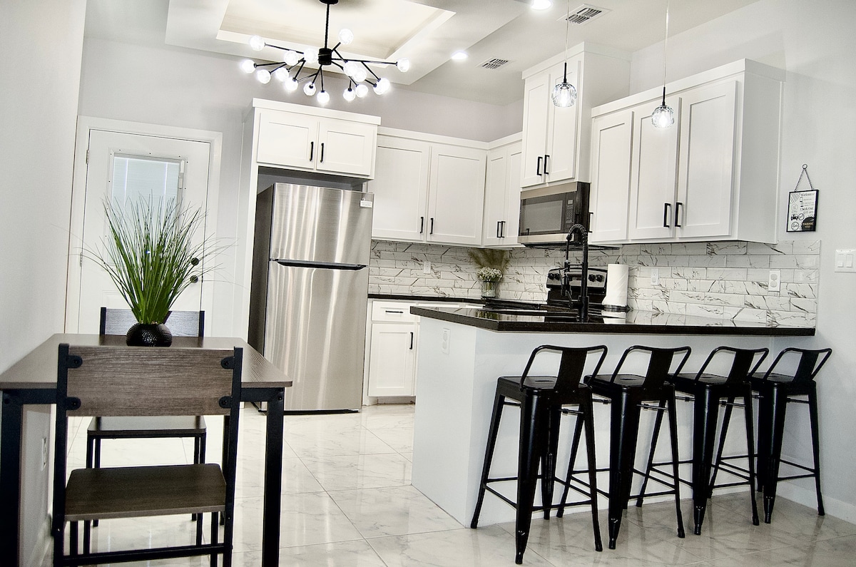 The kitchen features sleek white cabinetry and stainless steel appliances. A large island with seating is complemented by a dining table for four. Modern light fixtures illuminate the space, which showcases a clean marble backsplash and spacious countertops.